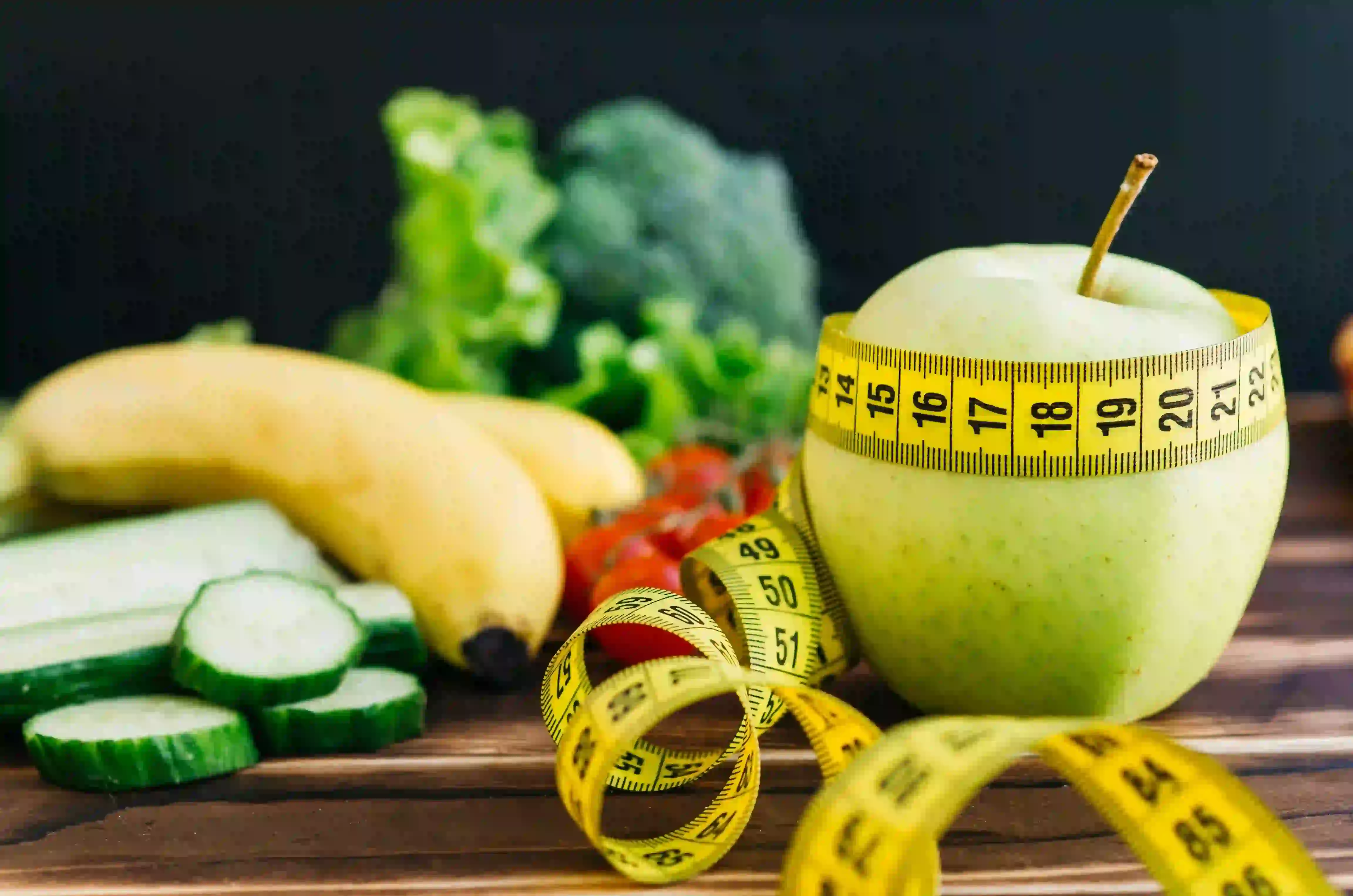 Healthy food including an apple, banana, cucumber, and broccoli with a yellow measuring tape