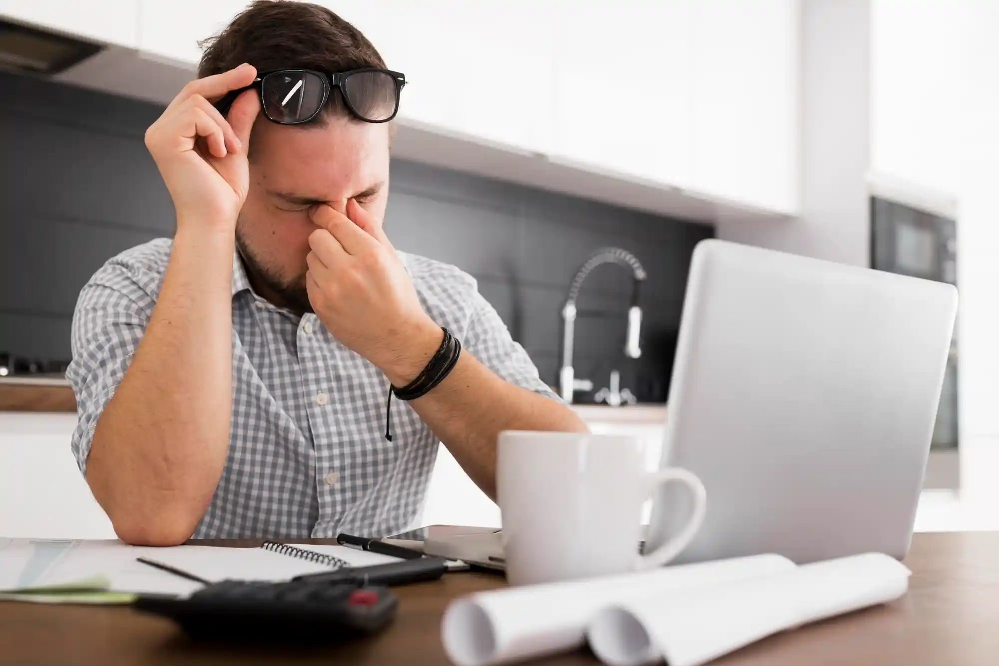 A man rubbing his eyes in pain while sitting in front of a laptop, suffering from digital eye strain