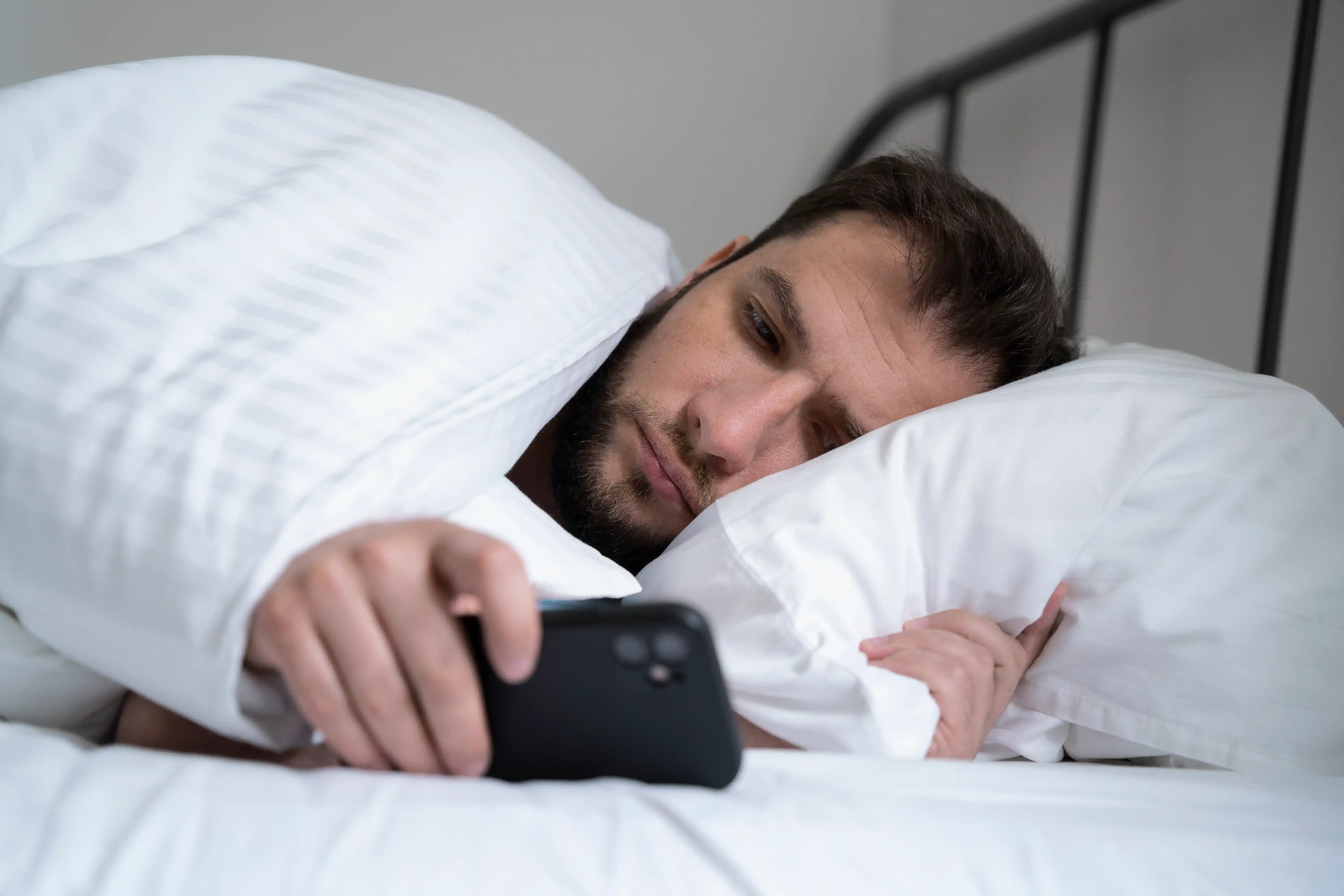 A man lying in bed looking tired and looking at his smartphone, illustrating sleep disruption